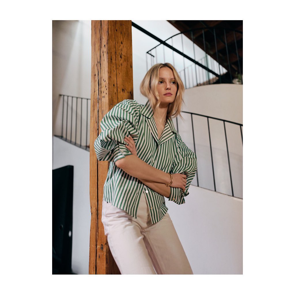Woman in green and white striped long-sleeve blouse leaning against a wooden beam inside a building with a staircase