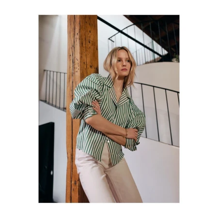 Woman in green and white striped long-sleeve blouse leaning against a wooden beam inside a building with a staircase