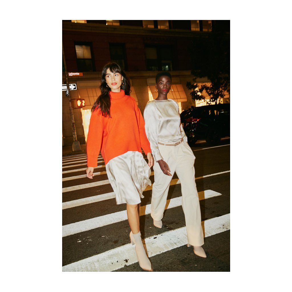 Two women in fashionable clothes walk on a zebra crossing in a city at night; one wears an orange sweater and the other a beige satin top.