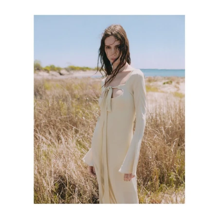 Model in a long cream cutout dress standing in tall coastal grass by the ocean under a clear sky.