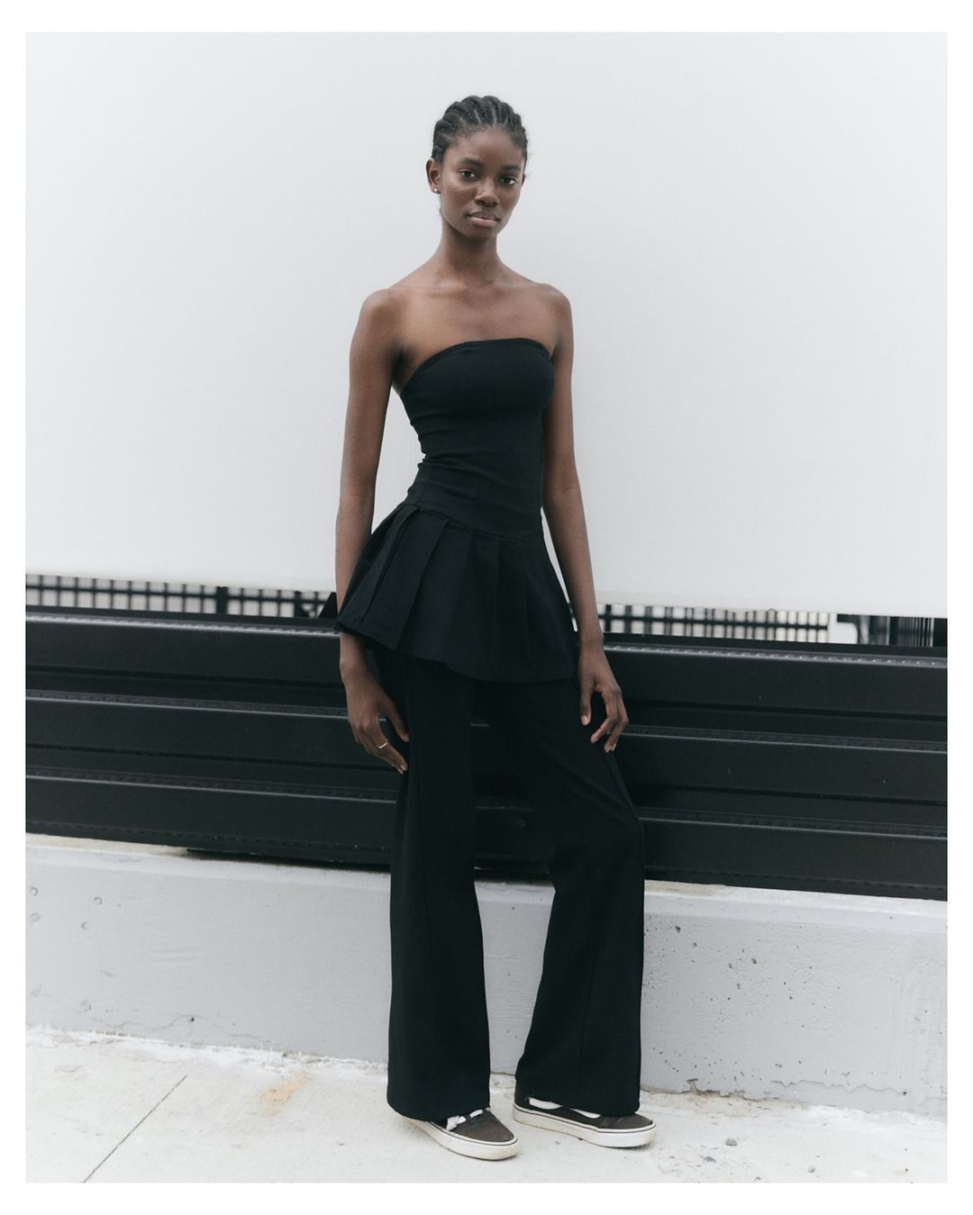 A Black woman model in a black strapless top and pants stands in front of a white wall with black horizontal bars. Fashion editorial.