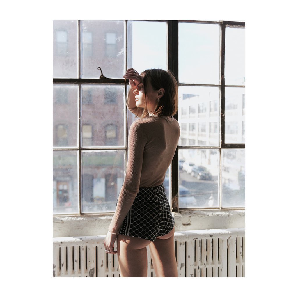 Young woman in shorts stands by a large window with a city view in soft lighting, styled in a minimalist aesthetic.