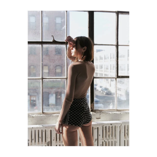 Young woman in shorts stands by a large window with a city view in soft lighting, styled in a minimalist aesthetic.