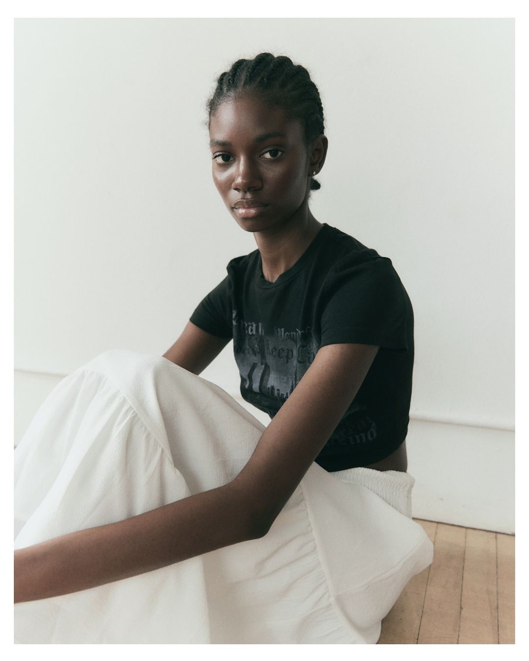 An African-American young woman with braided hair is sitting and wearing a black printed T-shirt and a long white skirt in the photo studio.