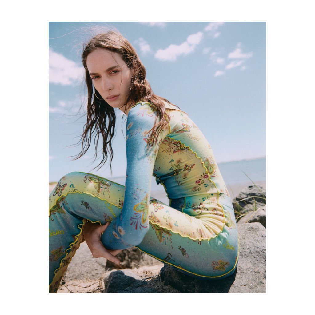 A young woman poses outdoors on a rocky beach in a blue-and-green patterned top and leggings, with the ocean and sky behind her.