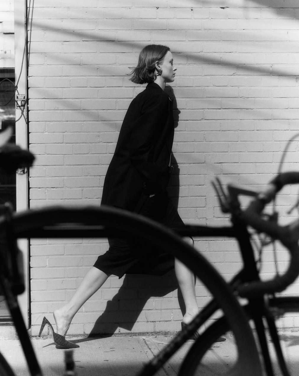 Black and white fashion shot: A female model in a dark coat walks past a brick wall, with a bicycle partially obscuring the foreground.