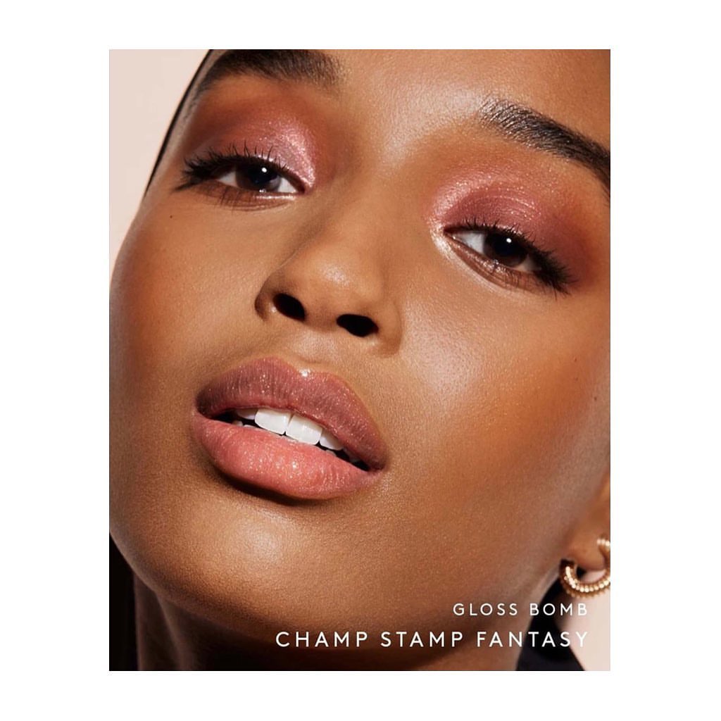 Close-up studio shot of a woman with dark skin and pink eyeshadow and lip gloss, featuring clean white teeth, and gold hoop earring.