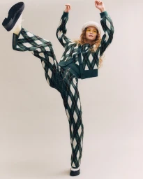 Model in green and white argyle cardigan and matching trousers kicking a leg up against a plain background