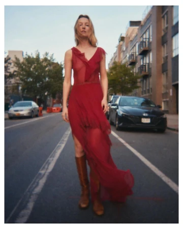 A woman in a long red dress and brown boots standing on an urban street with parked cars and buildings.