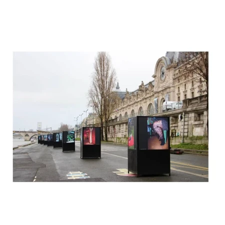 Outdoor art installation in Paris featuring several black display kiosks along the Seine River, showcasing various images against a skyline.