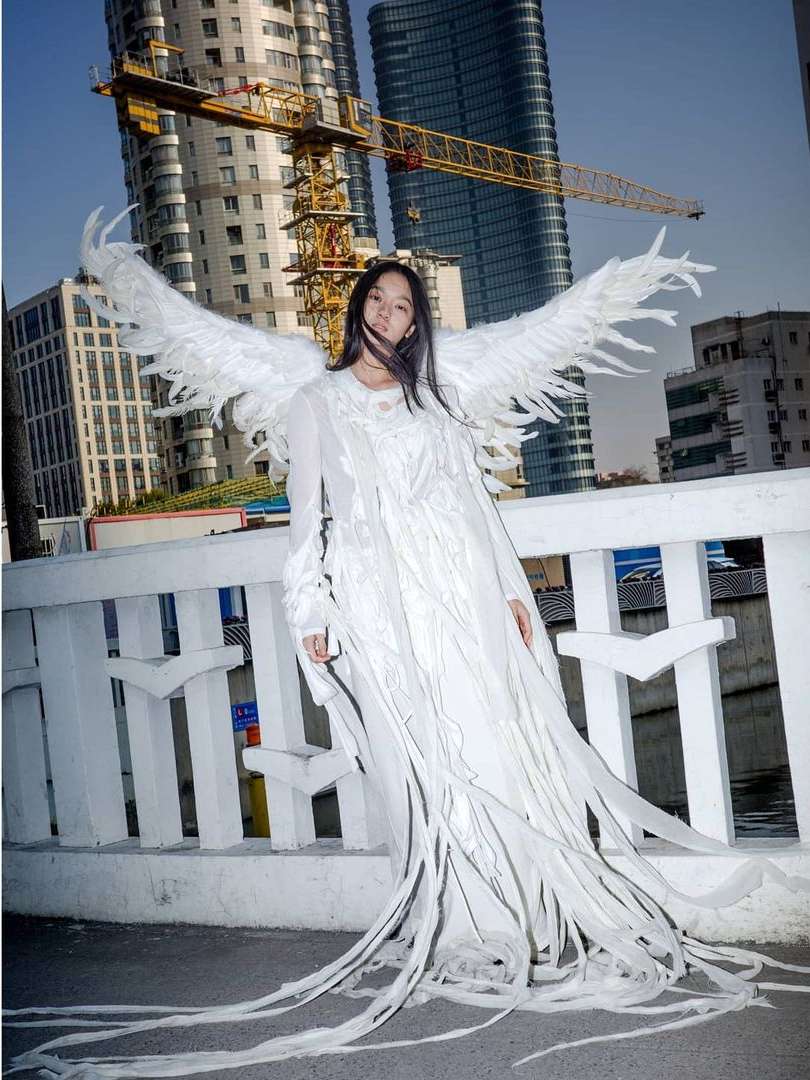 Woman in flowing white gown and angel wings poses in urban setting with modern buildings and construction crane in background.