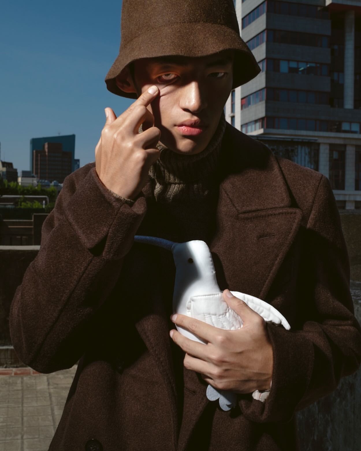 A young adult wears a brown coat and bucket hat, holding a plush white bird in an urban setting with clear sky and modern buildings.