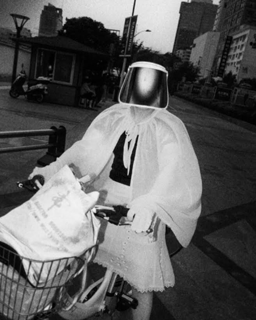 Person on bicycle wearing face shield and translucent poncho, with bag in basket, cityscape in background, black and white photograph.