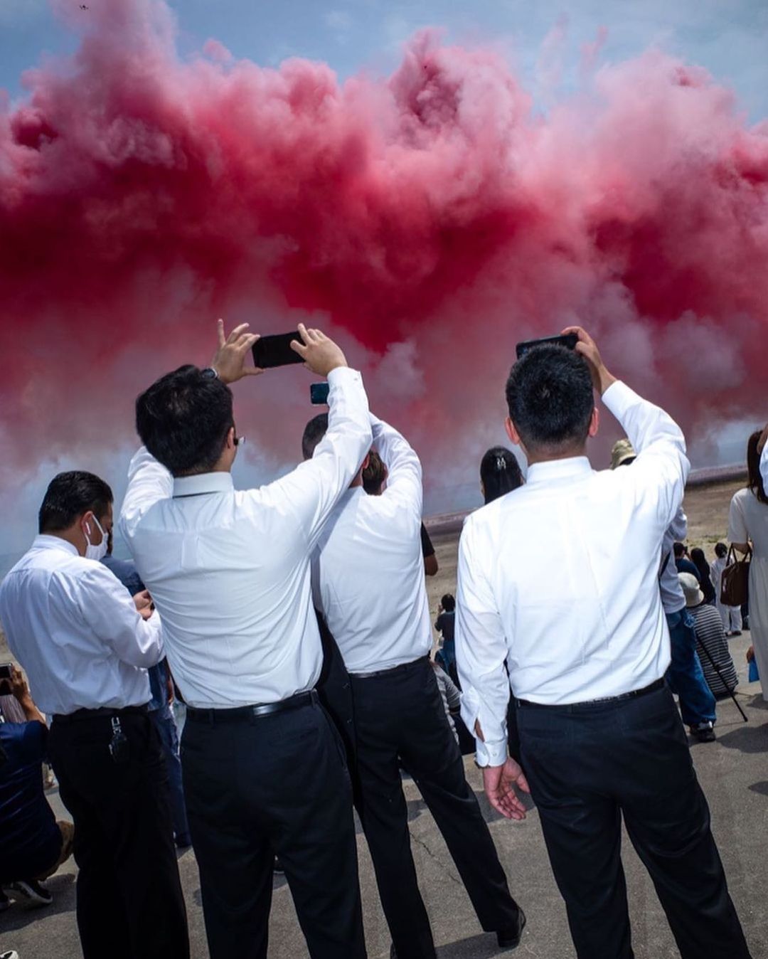A group of people photograph a red smoke display in an outdoor setting, dressed in white shirts and dark trousers, with blue sky.