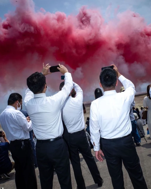 A group of people photograph a red smoke display in an outdoor setting, dressed in white shirts and dark trousers, with blue sky.
