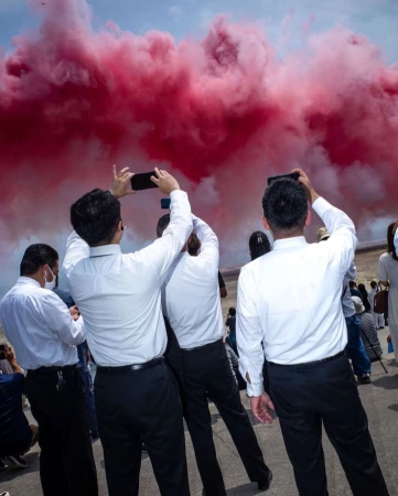 A group of people photograph a red smoke display in an outdoor setting, dressed in white shirts and dark trousers, with blue sky.