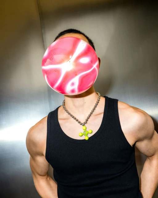 Asian male poses indoors with a pink and white patterned balloon obscuring his face, wearing a black tank top and a silver necklace.