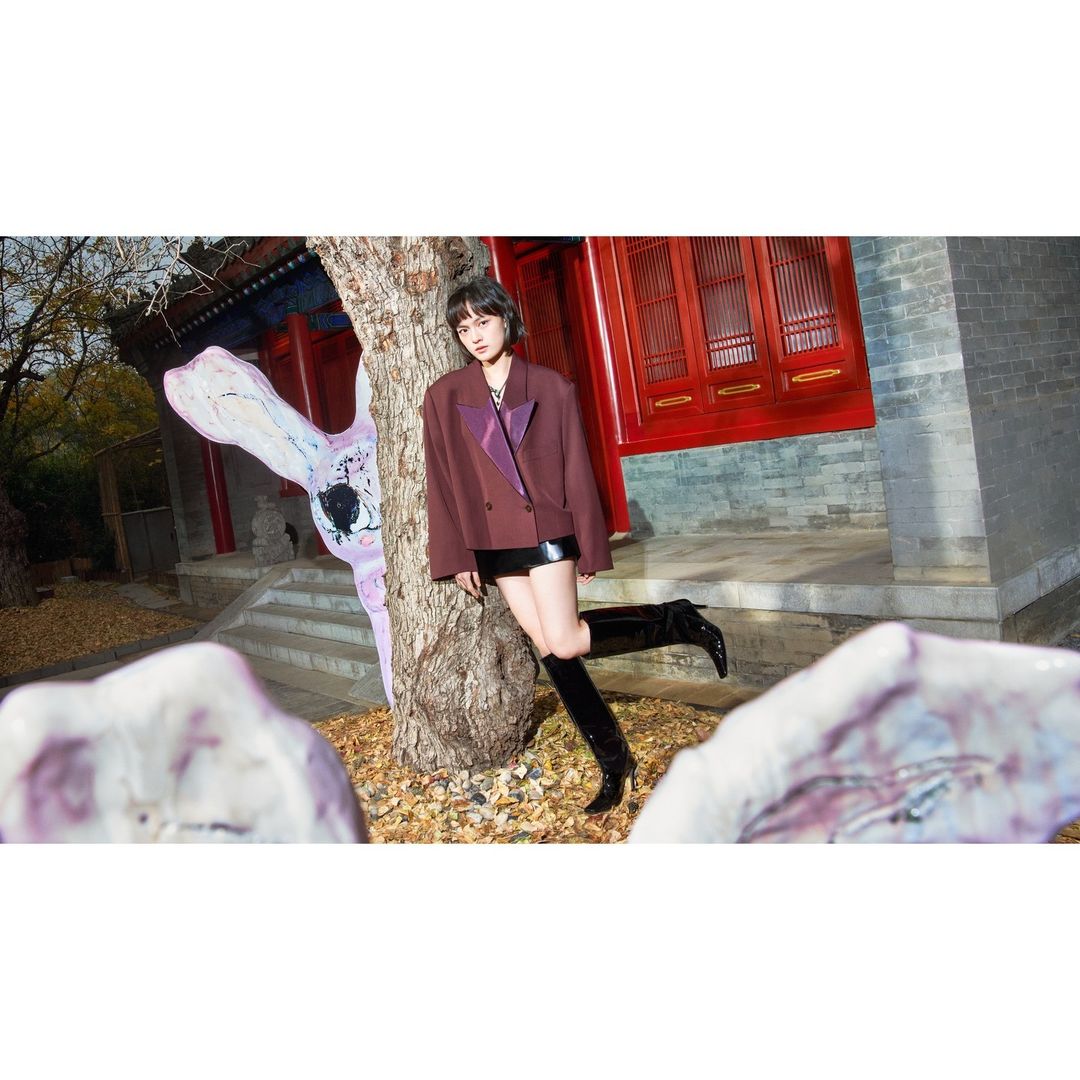 A young adult woman with short hair poses outdoors wearing a blazer and boots, leaning against a tree with a colorful building backdrop.
