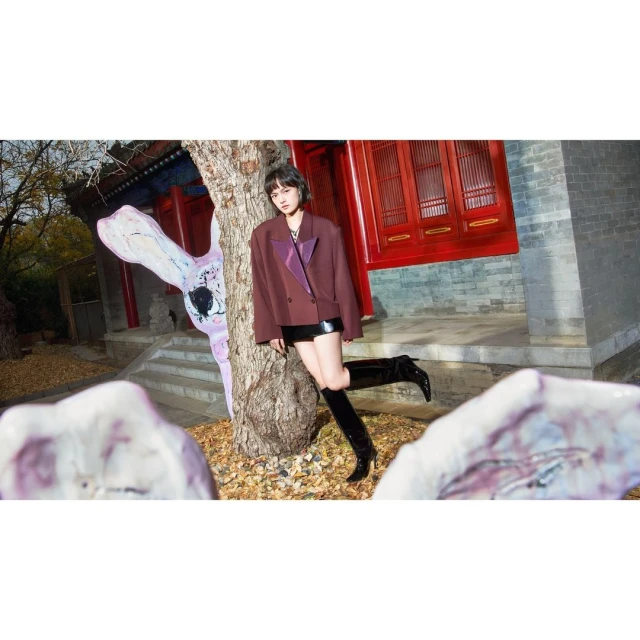 A young adult woman with short hair poses outdoors wearing a blazer and boots, leaning against a tree with a colorful building backdrop.