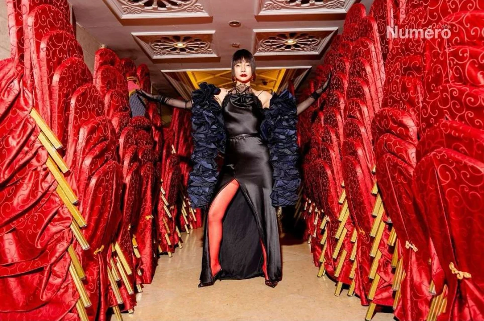 Fashion editorial image of a woman in a black gown posing amidst stacked red chairs, creating a striking visual corridor in a studio.