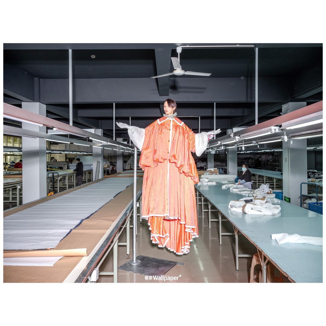 Model in a voluminous orange garment suspended in a bright garment factory with rows of sewing tables and sewing machines, editorial fashion.