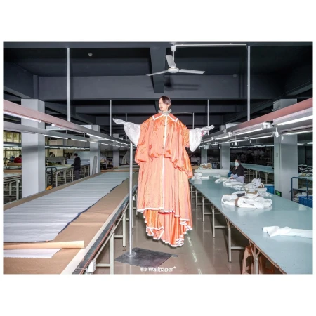 Model in a voluminous orange garment suspended in a bright garment factory with rows of sewing tables and sewing machines, editorial fashion.
