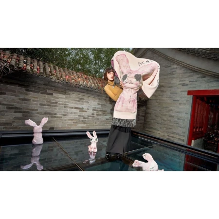 A woman poses outside wearing a rabbit-print scarf, next to rabbit figurines on a reflective surface, with a brick building backdrop.