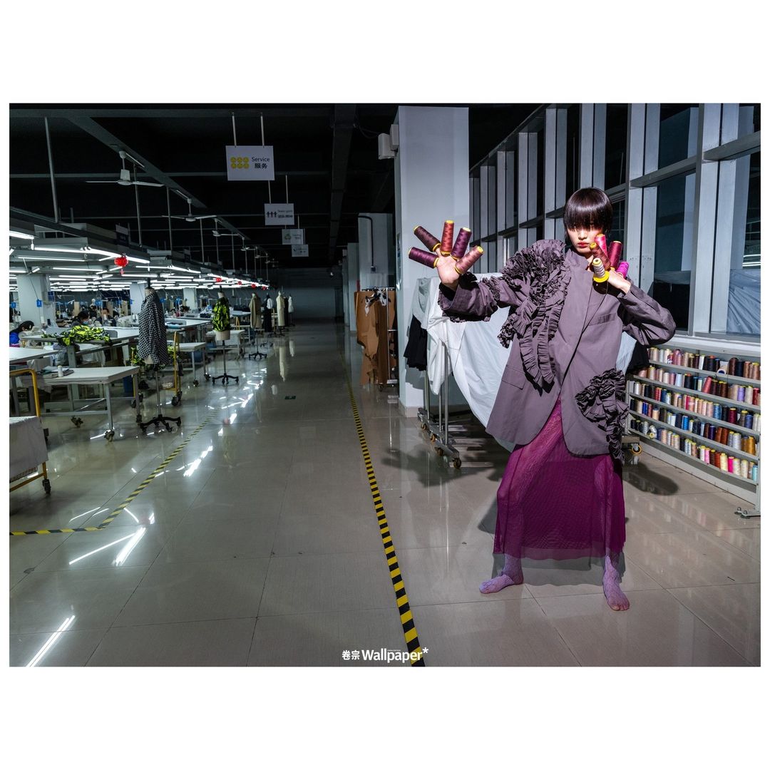 Adult female poses in a purple dress and blazer jacket in a textile factory holding thread spools, rows of sewing machines in background.
