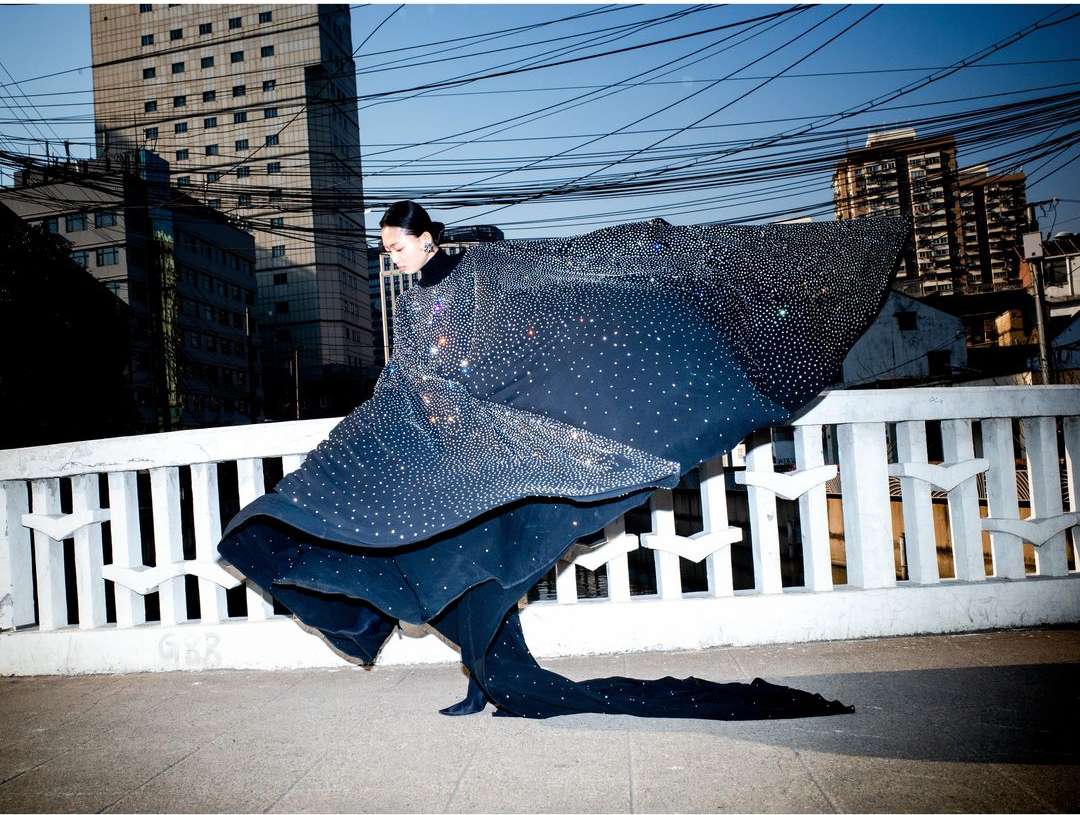 Woman in a glittering gown with a train poses on a bridge with an urban background, showcasing a blend of fashion and city life.