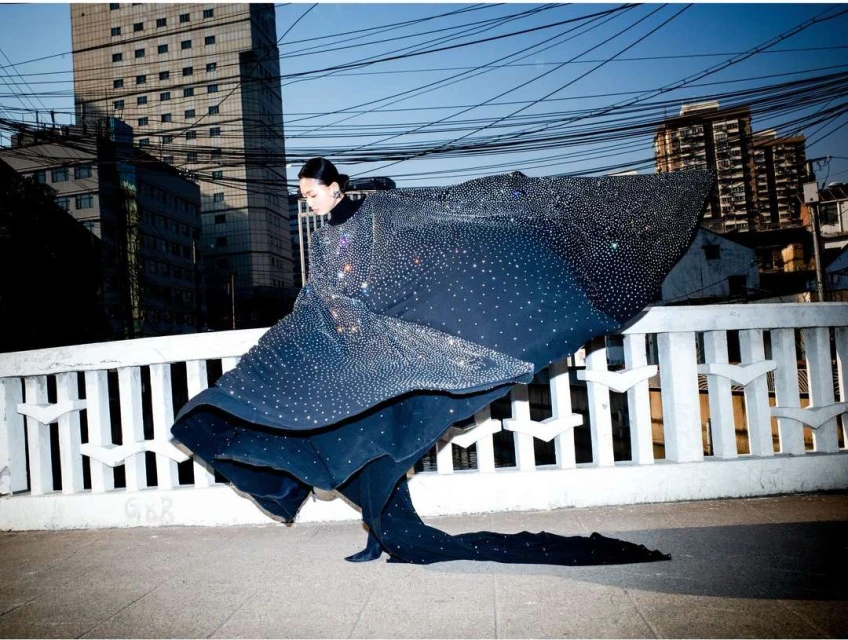 Woman in a glittering gown with a train poses on a bridge with an urban background, showcasing a blend of fashion and city life.