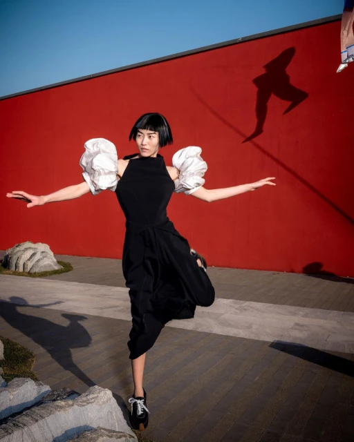 Woman in black dress with white puff sleeves strikes a ballet pose in an outdoor garden setting with a red wall backdrop on a sunny day.