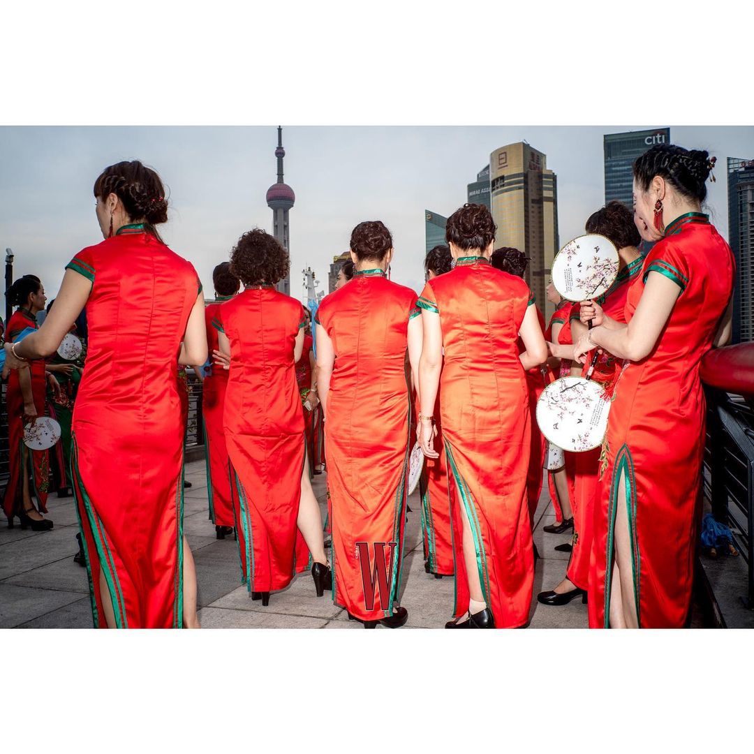 A group of women in traditional red Chinese dresses stand in an urban setting, with a distinctive tower and modern buildings in the background.