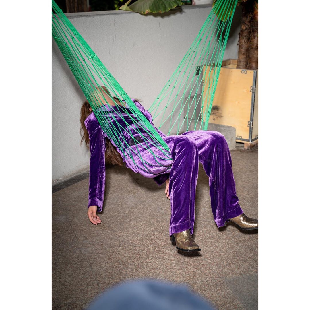 Adult female in a purple velvet suit relaxes in a green woven hammock, with muted gold cowboy boots visible, against an outdoor wall.