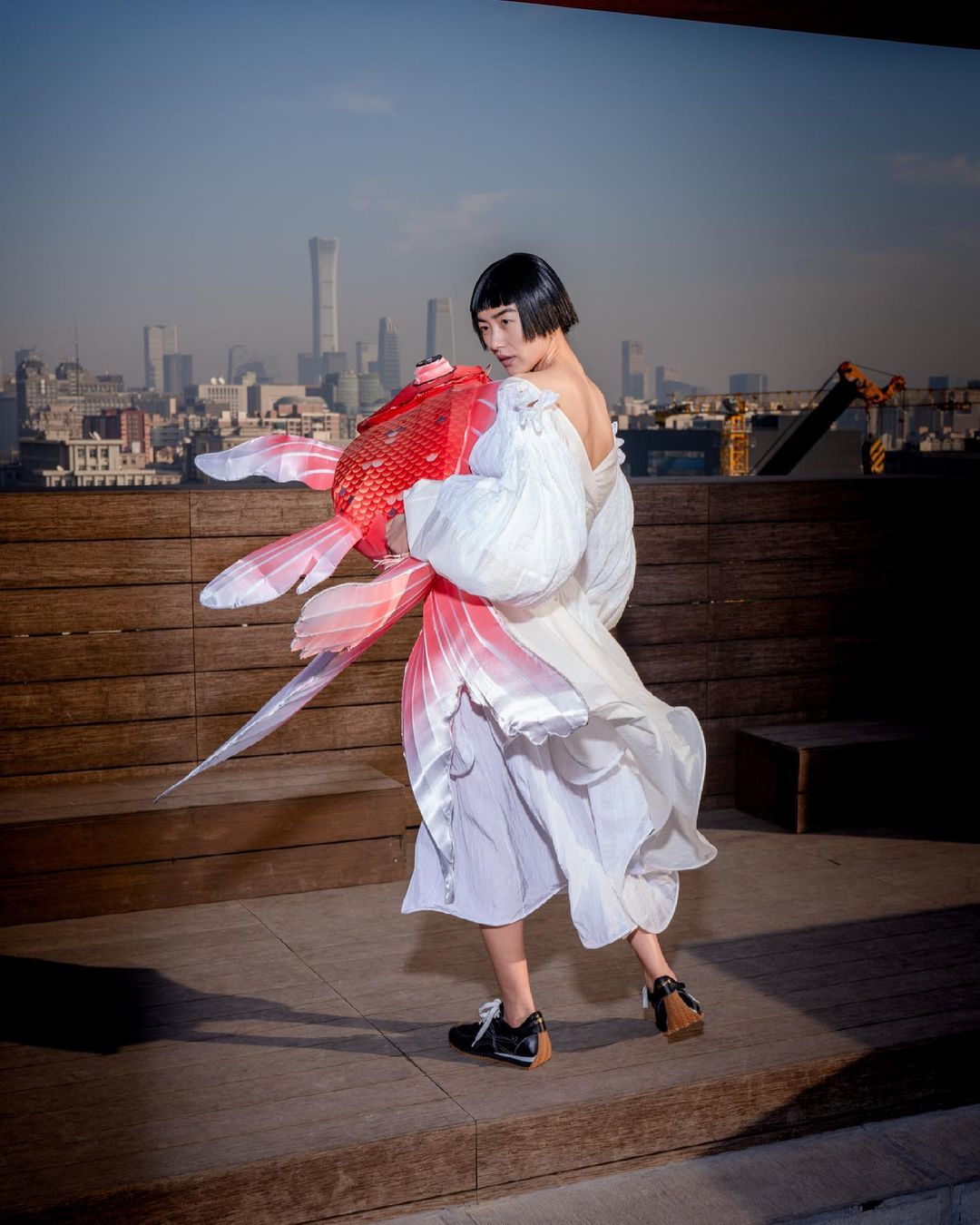 Woman in a white dress holds a large red and white origami goldfish with a cityscape background on a rooftop in the daytime.