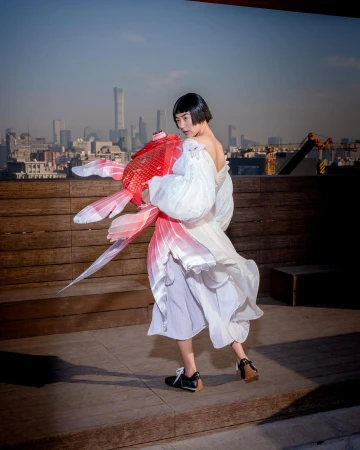 Woman in a white dress holds a large red and white origami goldfish with a cityscape background on a rooftop in the daytime.
