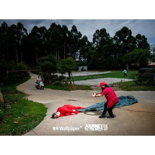 Outdoor staged photo of a person lying on a sheet while another person pulls the sheet in a park-like setting with trees and a scooter.