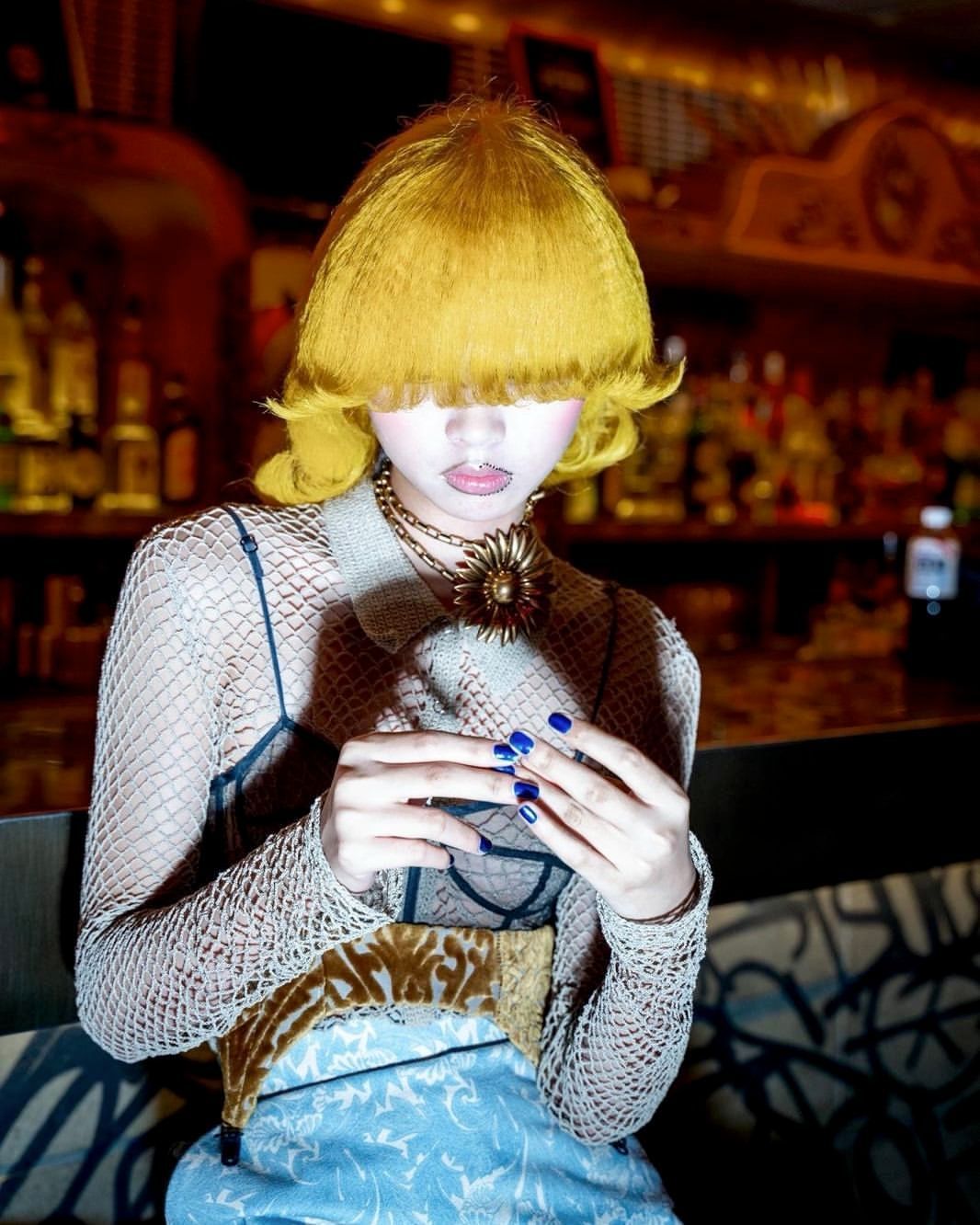 Indoor portrait of a young adult with yellow hair, a fishnet top, a large necklace, and blue manicured nails against a dark bar setting.