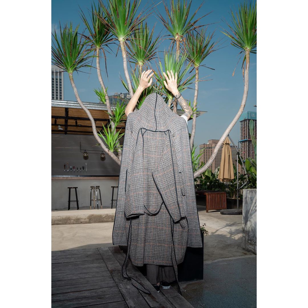 A person wearing a grey, plaid hooded coat outdoors, standing behind a palm tree with a building visible in the background on a clear day.