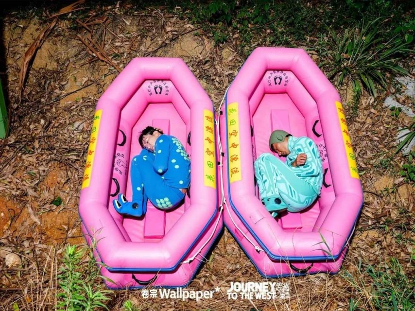 Two adults rest in pink inflatable rafts outdoors. One wears a blue polka-dot outfit, the other wears a pale green patterned suit.