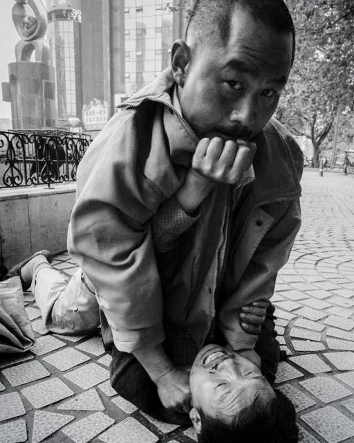 Black and white portrait of a man standing over another man lying on a patterned tile surface in an outdoor urban environment.