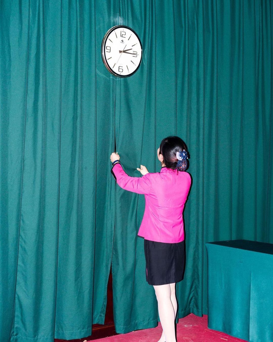 Woman in pink jacket adjusts string attached to wall clock against teal curtain backdrop. Person reaching, time-telling concept.