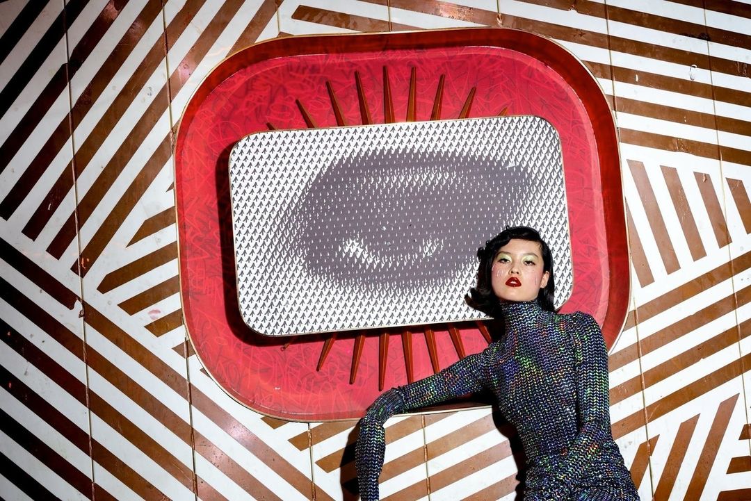 Portrait of a woman in a sparkly top posing in front of a red retro frame and brown and white striped background.