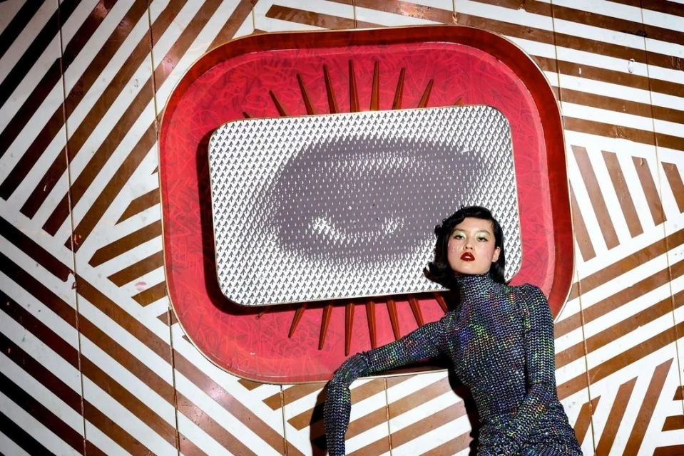 Portrait of a woman in a sparkly top posing in front of a red retro frame and brown and white striped background.