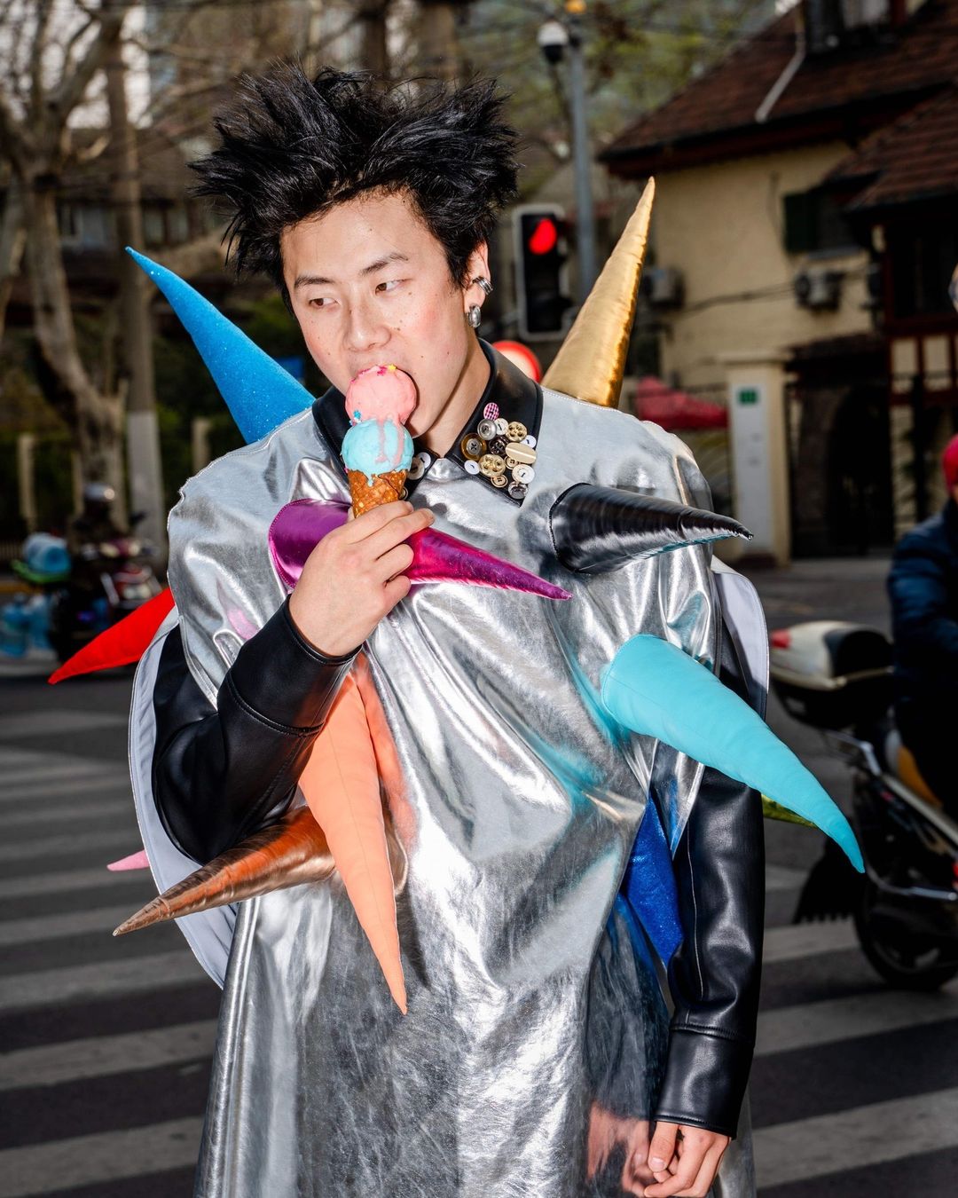 A person with spiky black hair stands on a street eating an ice cream cone, wearing a futuristic silver jacket with colorful cone shapes.
