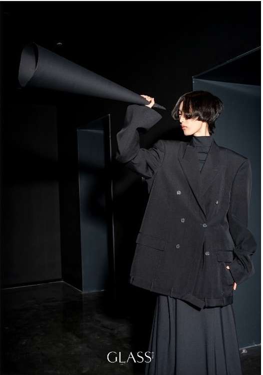 Stylish adult poses indoors with an oversized black paper cone, wearing a black blazer and skirt; a minimalist fashion shot.