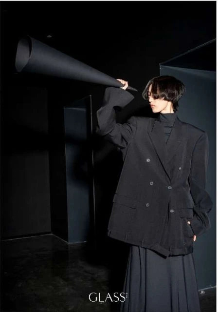 Stylish adult poses indoors with an oversized black paper cone, wearing a black blazer and skirt; a minimalist fashion shot.