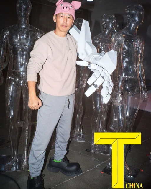 Adult Asian man with a pig hat and robotic arm stands in front of transparent mannequins in a photo studio. Fashion editorial.
