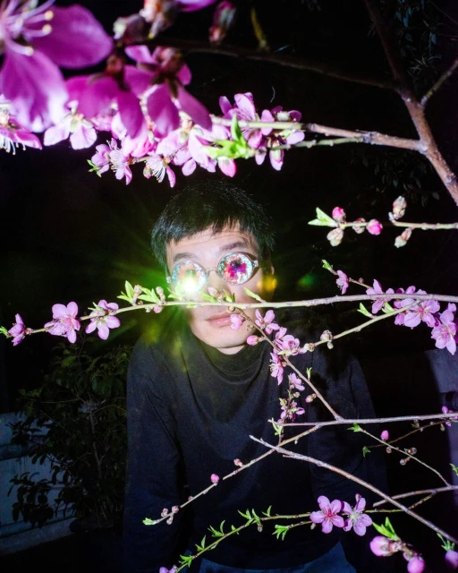 An individual wearing glasses with pink flowers reflected in the lenses, standing amidst branches of pink flowers in a dark outdoor setting.