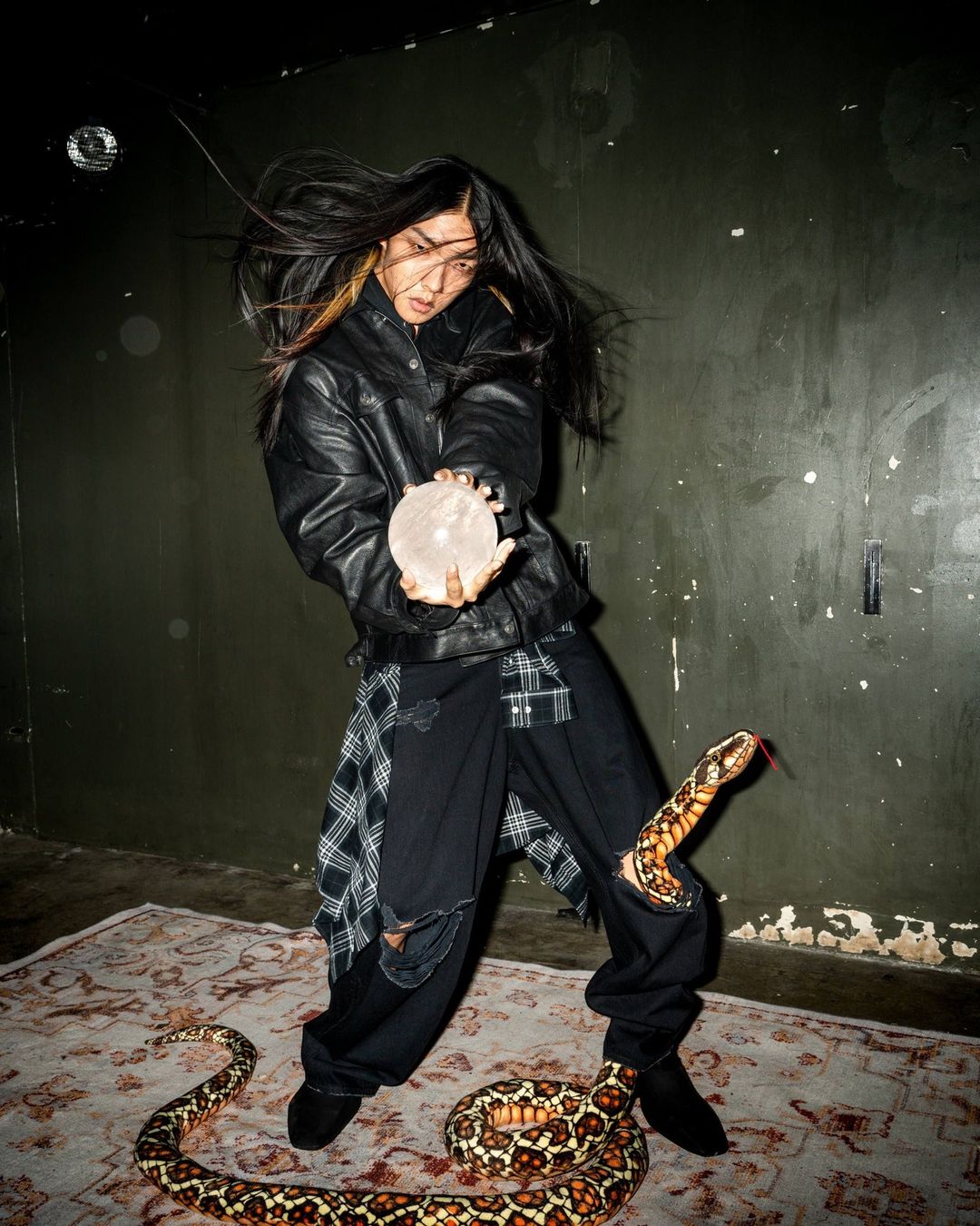 An Asian woman poses with a crystal ball and snakes in an edgy fashion shot set indoors with a patterned rug and a green backdrop.