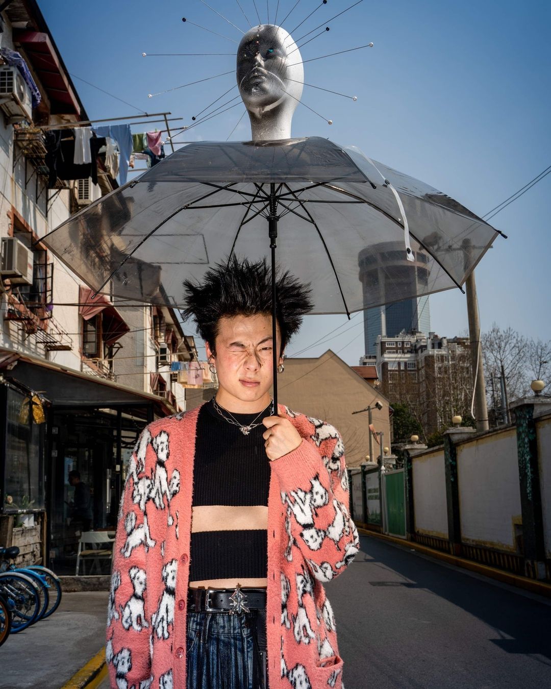 A young adult in a patterned cardigan stands on a street under an umbrella with a mannequin head hovering above him, creating a surreal scene.
